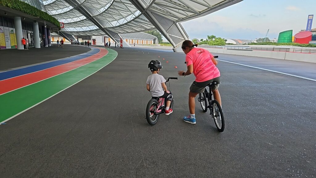 A Saddlewerkz coach riding beside a small helmeted learner through a cone drill under the National Stadium roof