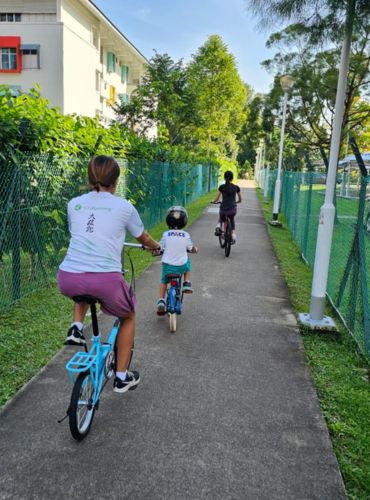 A coach on a blue bike riding beside a very small child on a balance-sized bike, with another learner ahead, on a park path